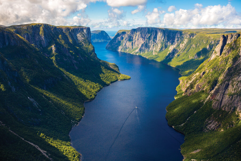 Aerial view of a deep blue fjord surrounded by steep, green-clad cliffs under a partly cloudy sky.
