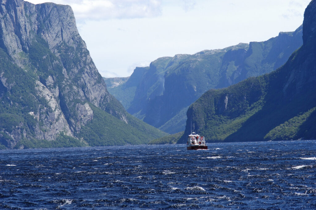 A boat sails on a large body of water surrounded by towering, rugged mountains covered in greenery under a partly cloudy sky.
