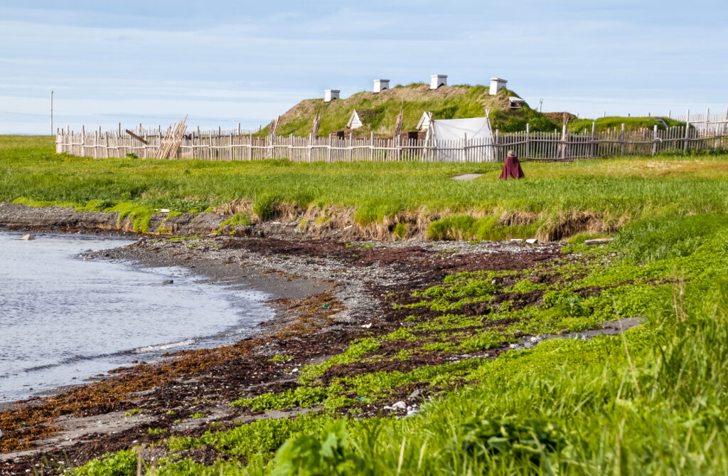 A coastal scene with a grassy shoreline, a wooden fence enclosing turf-roofed buildings in the background, and a person in a red cloak walking near the fence.