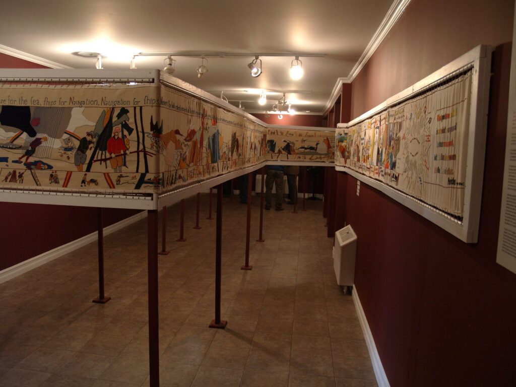 A long, detailed tapestry displayed in a narrow, well-lit room with maroon walls and tiled floor, depicting various historical scenes and figures.