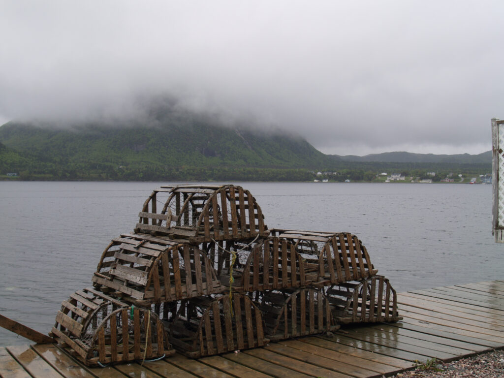 A stack of wooden lobster traps on a wet dock with a misty mountain and calm water in the background.