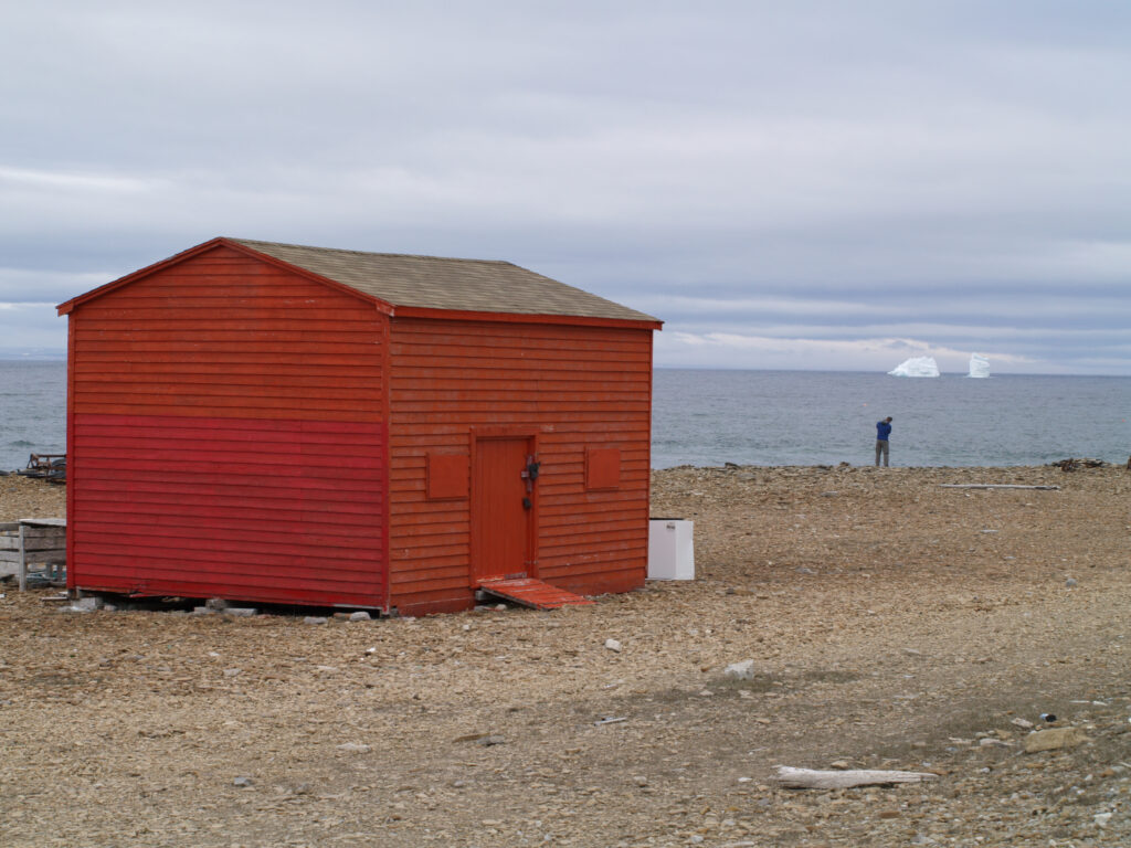 A red wooden shed stands on a rocky shore with a person in the distance taking a photo of two icebergs floating in the ocean under a cloudy sky.