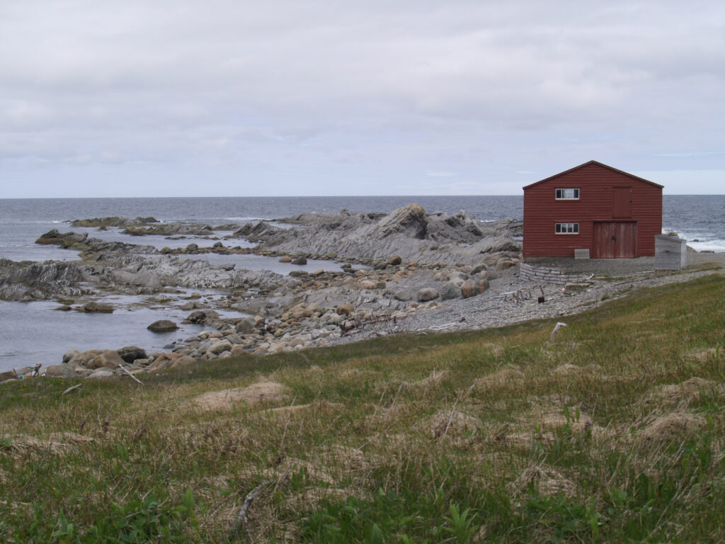 A red wooden building stands on a rocky shoreline with the ocean and jagged rocks extending into the water under a cloudy sky.