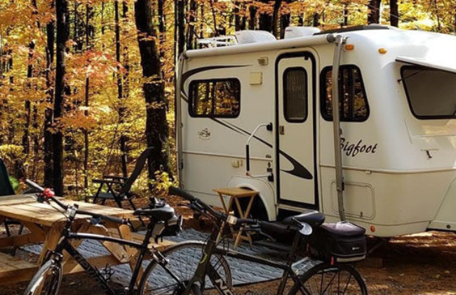 A white camping trailer parked in a forest with autumn foliage, accompanied by two bicycles and a picnic table.