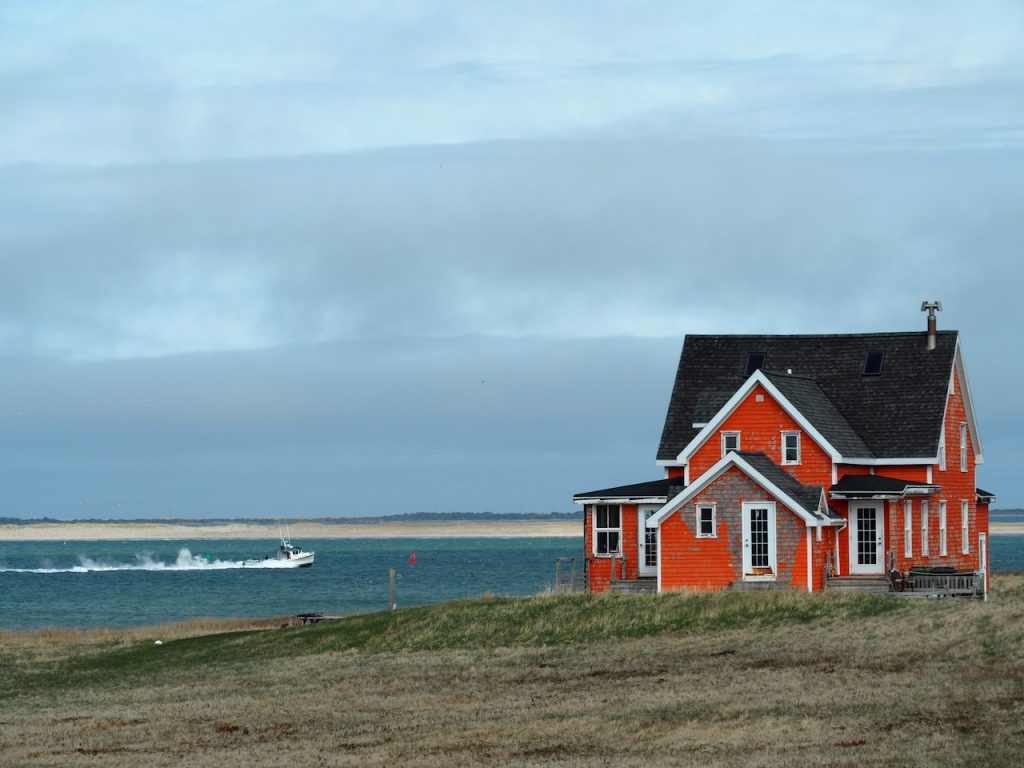 A bright orange wooden house sits on grassy land beside the water, with a small boat speeding past in the background off the Magdalen Islands.