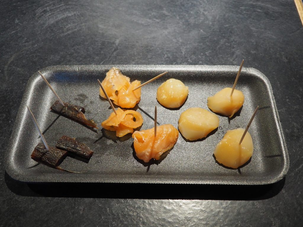 A black tray displays samples of smoked herring, mackerel, salmon, and scallops, each pierced with a toothpick for tasting.
