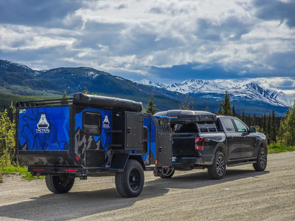 A black pickup truck is seen pulling a Tactical Overland trailer in the wilderness under a bright but cloudy sky.