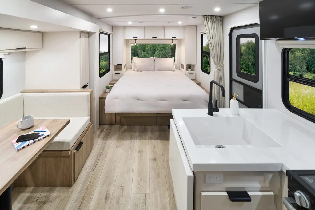 Interior of the 2026 Winnebago Thrive travel trailer featuring a modern kitchen with a white countertop, a dinette with light wood accents, and a convertible sofa bed against the back wall.