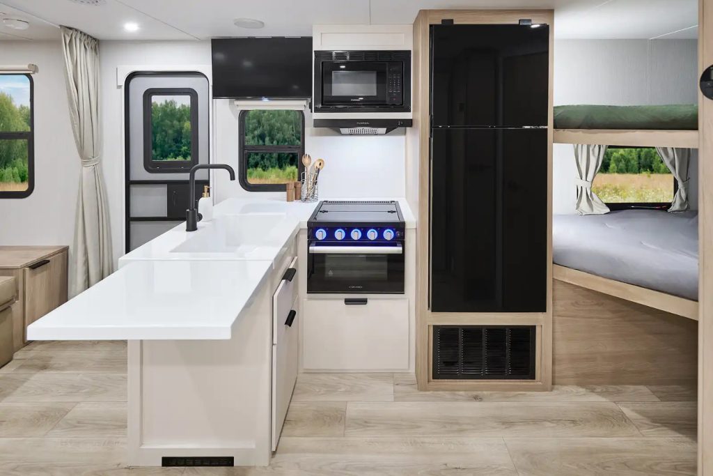 Interior view of a Winnebago Thrive travel trailer kitchen featuring a white island with sink, black refrigerator, microwave, stove with blue-lit knobs, and bunk beds in the background.