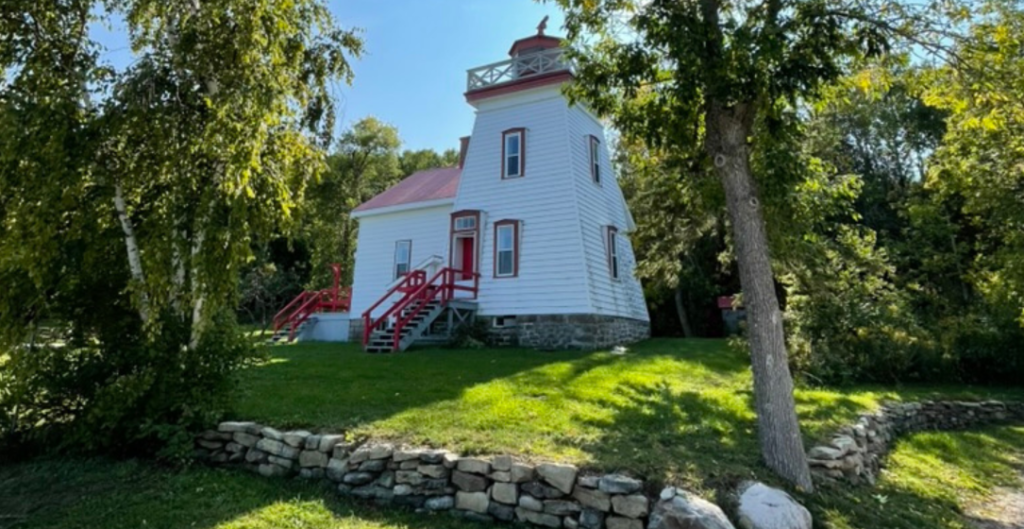 A white, two-story lighthouse with red trim and a red roof stands on a grassy lawn surrounded by trees.