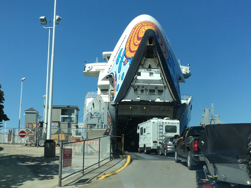 Vehicles, including an RV, drive onto the MS Chi-Cheemaun ferry with a colorful painted bow under a clear blue sky.