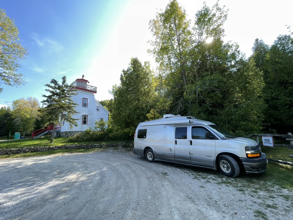 A silver camper van is parked on a gravel lot near the Janet Head lighthouse surrounded by green trees under a bright sky.