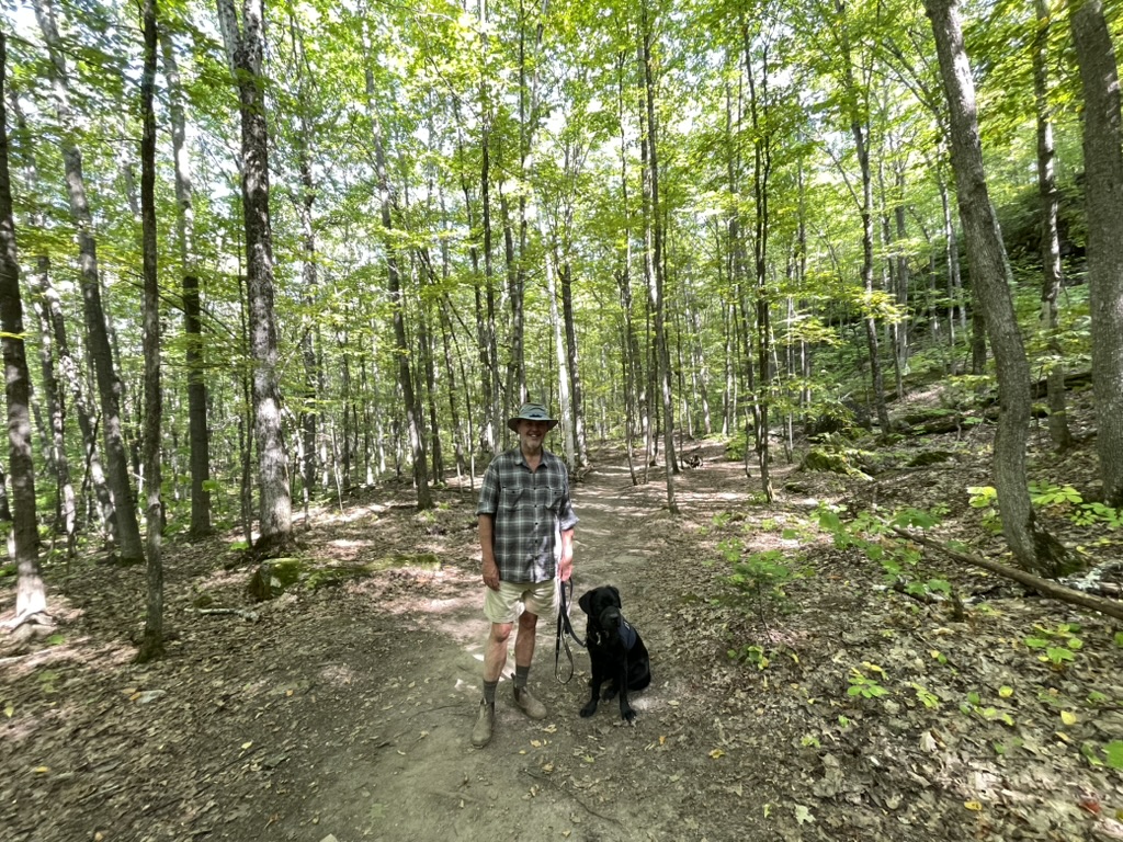 A person wearing a hat stands on the Cup and Saucer Michigiwadinong Hiking Trail with a black dog on a leash, surrounded by tall green trees.