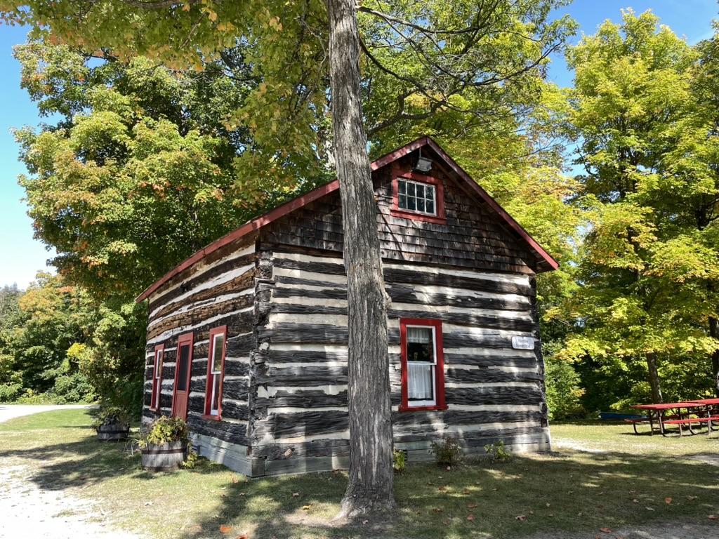 A rustic log cabin with red-trimmed windows stands among green trees on a sunny day at the Centennial Museum.