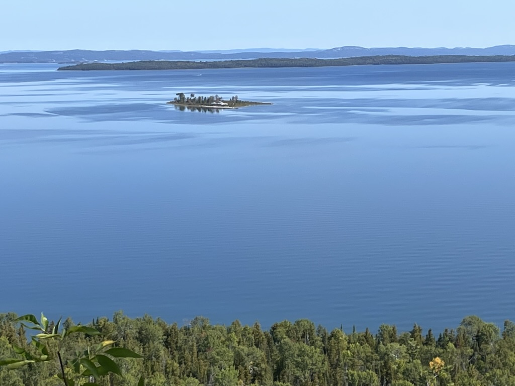 A small tree-covered island sits in the middle of a calm blue lake on Sheguiandah Bay, surrounded by distant forested shores under a clear sky.