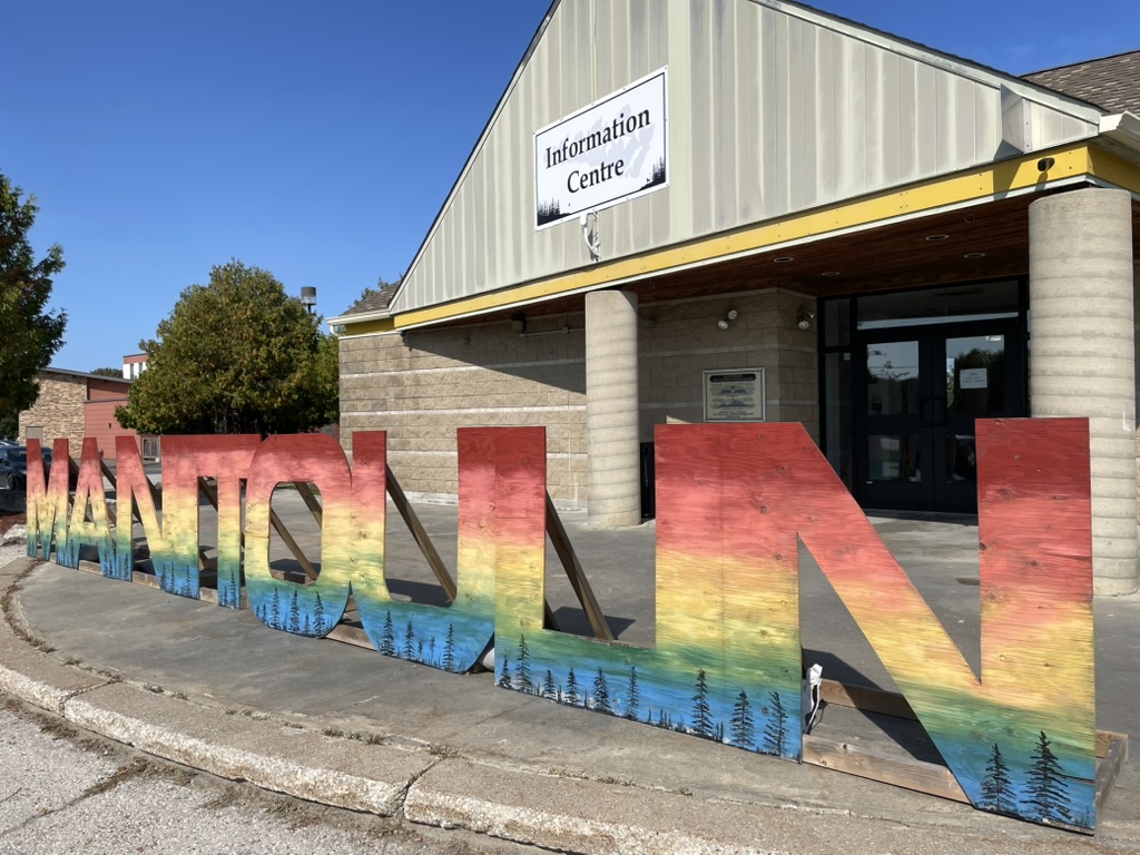Large wooden letters spelling “MANITOULIN,” painted in rainbow colors with tree silhouettes, stand in front of an Information Centre building.