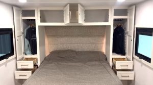 Interior view of an RV bedroom featuring a large bed with gray bedding, overhead storage cabinets with doors open, and two side wardrobes with hanging clothes and drawers partially open.