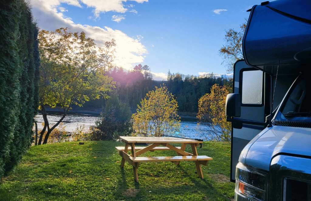 An RV is parked on a campsite next to a lake and the sun is rising over the lake.