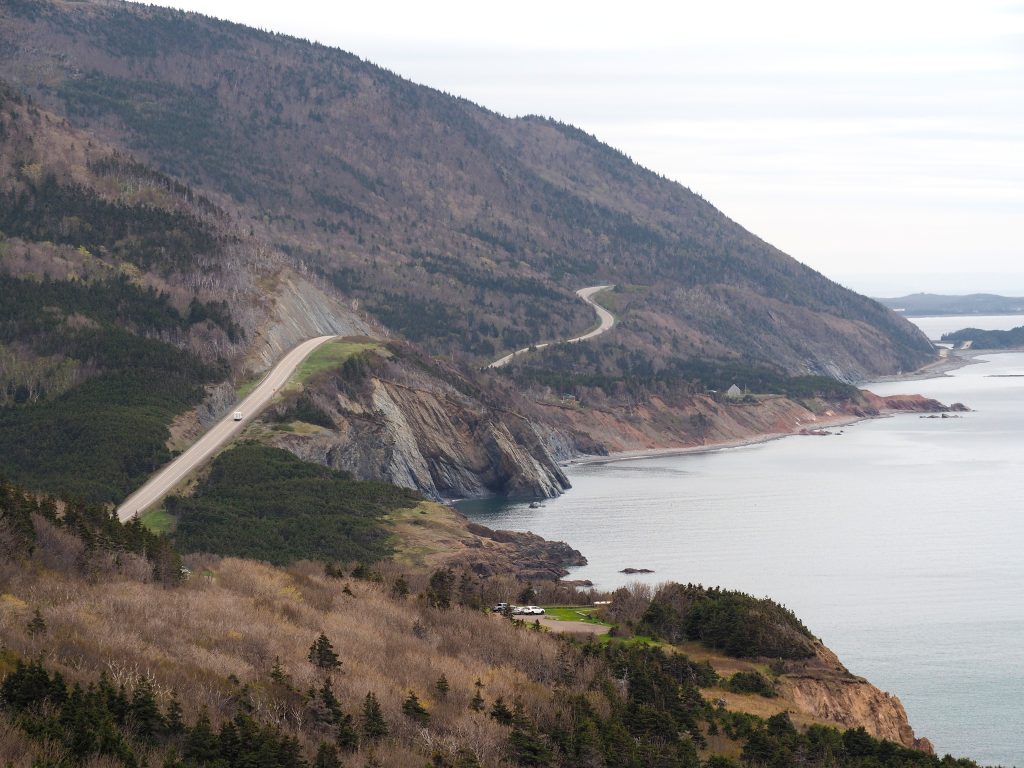A winding coastal highway along steep cliffs overlooking the ocean, with forested hills and rocky shoreline stretching into the distance.