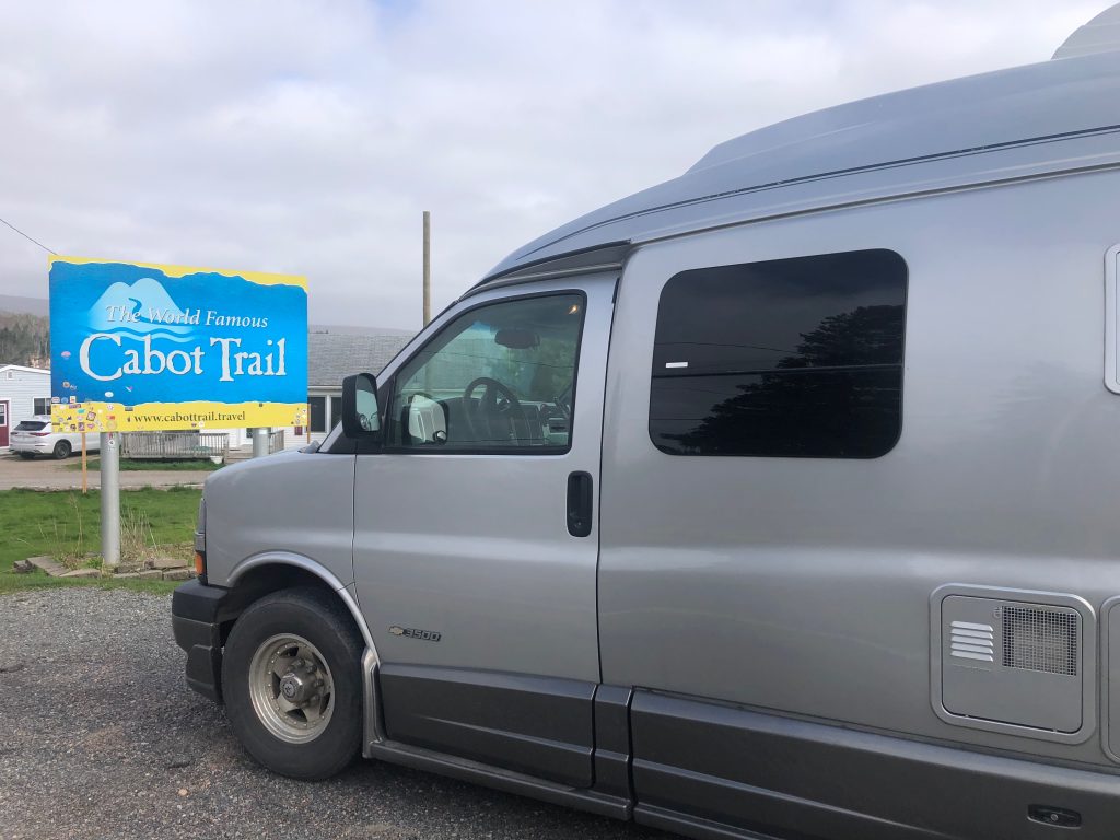 A silver camper van parked near a blue roadside sign that reads ‘Cabot Trail,’ with buildings, grass, and a cloudy sky in the background.