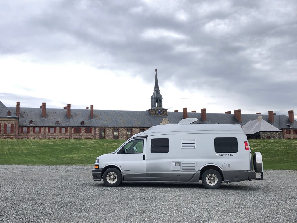 A silver camper van parked on a gravel area in front of a long historic building with red‑trimmed windows, stone walls, and a central clock tower under an overcast sky.
