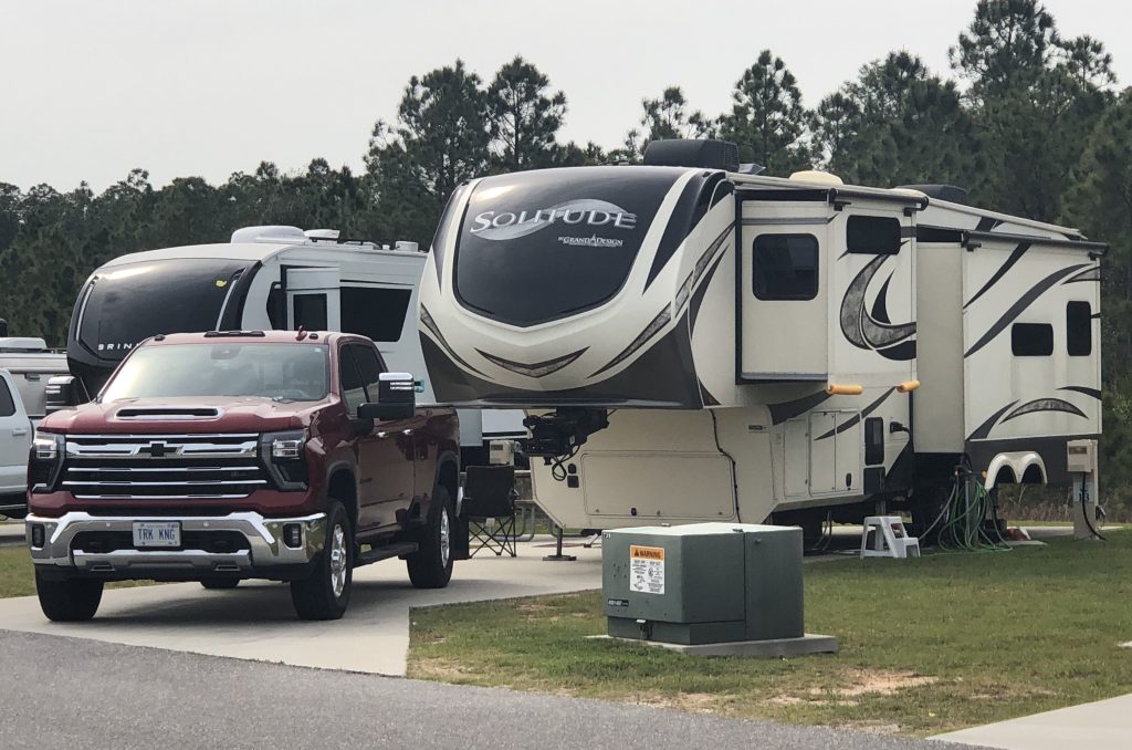 A beige and black fifth‑wheel RV parked at a campsite beside a red pickup truck, with additional RVs and a line of tall pine trees in the background.