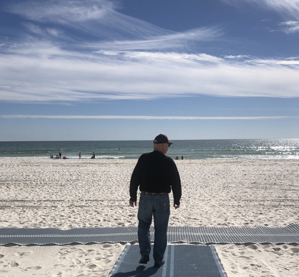A person standing on a beach access mat facing the shoreline, with white sand, gentle waves, and a blue sky with wispy clouds.