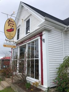 The exterior of the Red Shoe Pub, a white wooden building with large front windows, red trim, and a hanging sign featuring a red shoe, with shrubs and steps leading to the entrance.