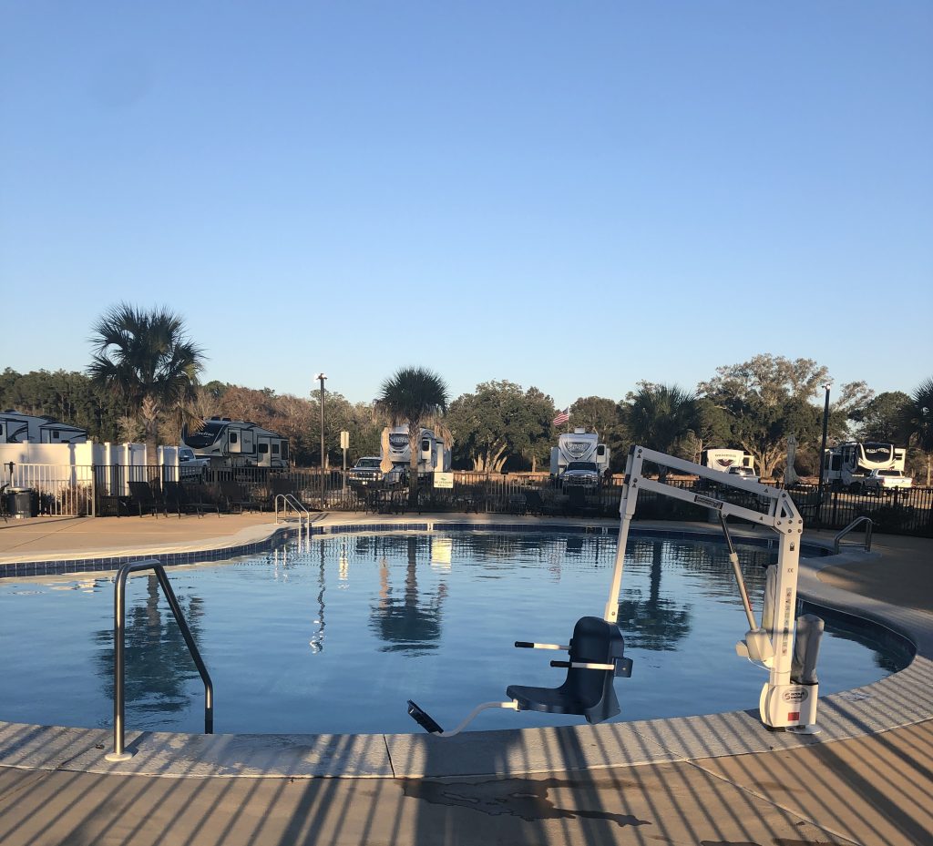 A calm outdoor swimming pool with accessibility equipment, surrounded by RVs, palm trees, and clear blue sky.