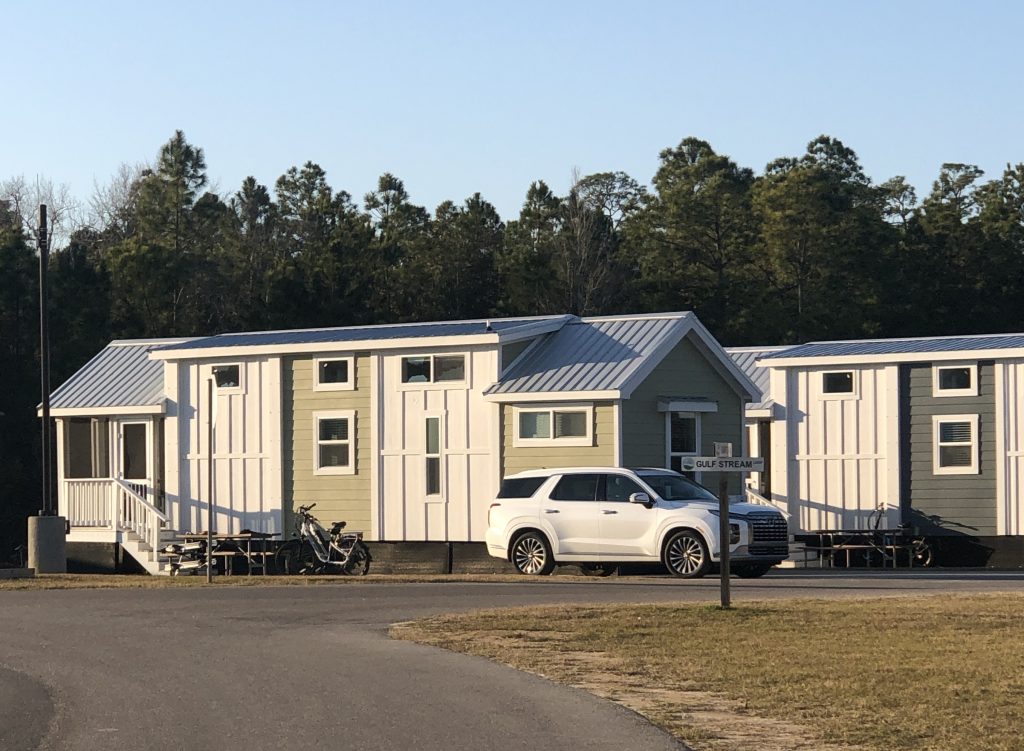 A small cottage-style tiny home with light siding and a metal roof, parked beside a white SUV, with additional tiny homes and trees in the background.