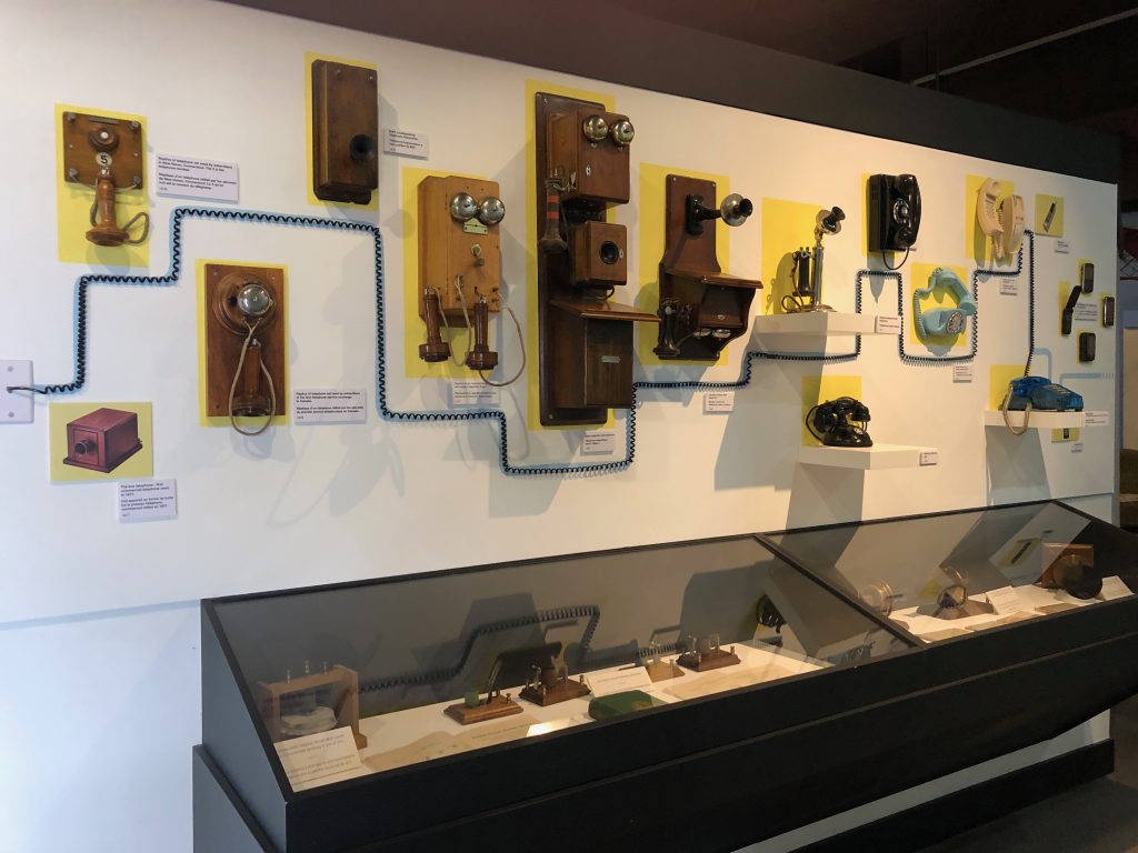 A museum exhibit featuring a wall display of vintage telephones arranged in a timeline‑style layout, with various wooden and plastic phone models mounted above a glass case containing additional communication artifacts.