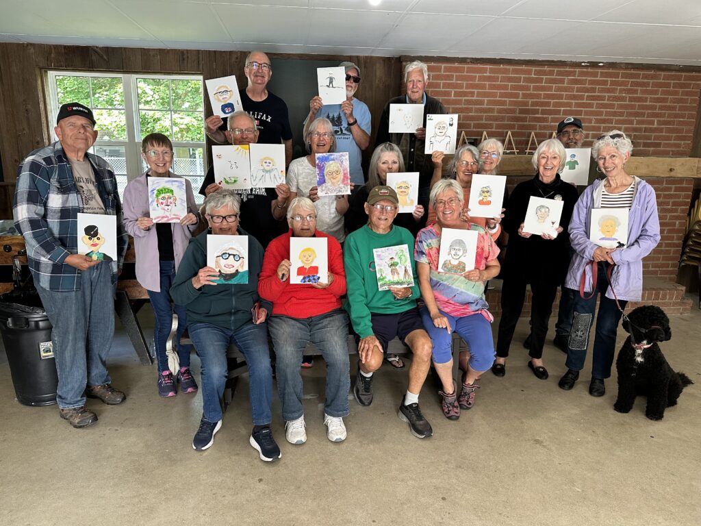 Group of RV club members indoors displaying self-portrait paintings created during a campground portrait art challenge, with a black dog seated at the front.