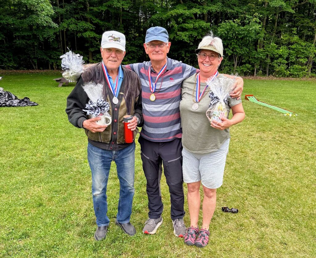 Three RV club participants wearing medals and holding prize baskets after placing in an Olympic-style campground competition held on a grassy field.