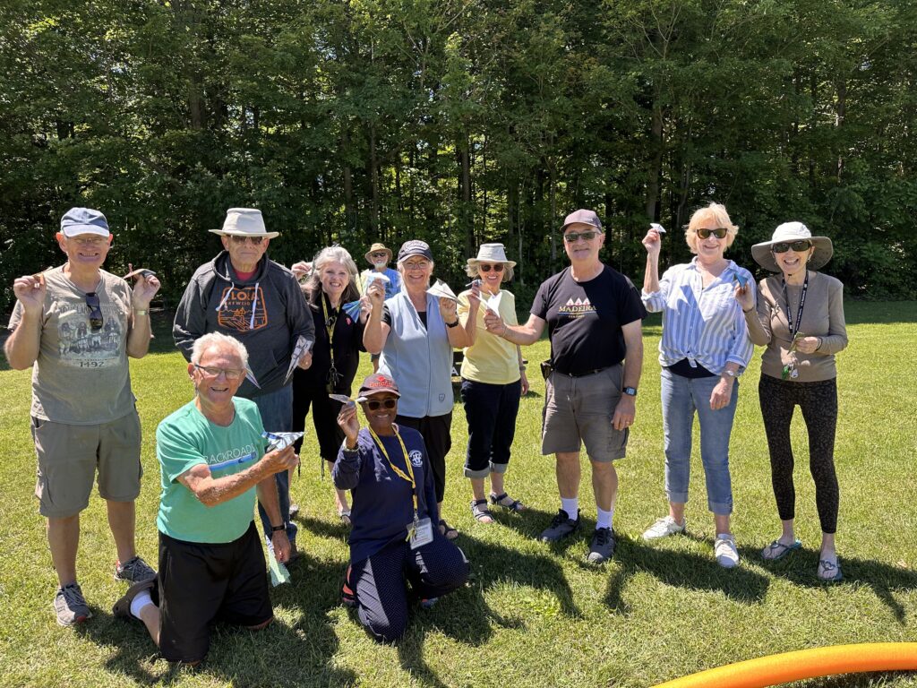 Group of RV club members outdoors displaying paper airplanes during a paper airplane design and flying competition on a sunny campground lawn.