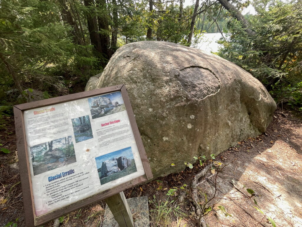 An interpretive panel with information on a trail at Finlayson Point Provincial Park next to a large glacial boulder.