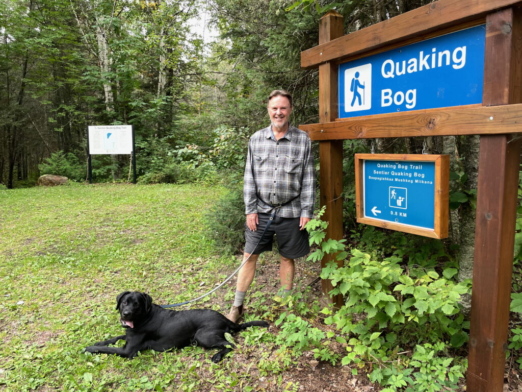A person stands beside a Quaking Bog trail sign at Ivanhoe Lake Provincial Park with a black lab on a leash.
