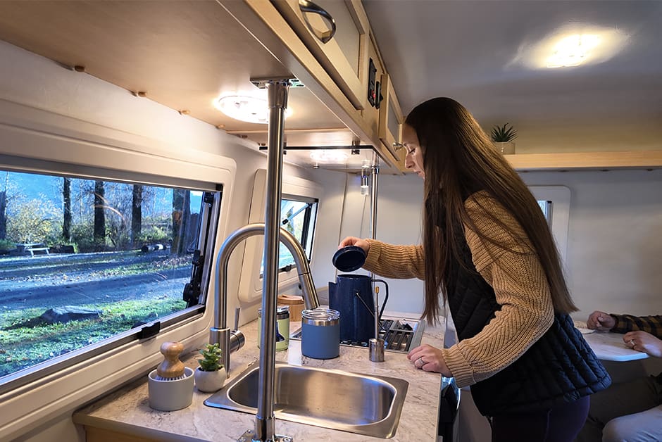 Young adult in the kitchen of the Escape E13 travel trailer, standing in front of the sink and cooktop.