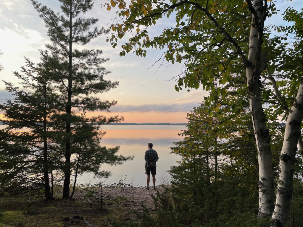 A person standing at the edge of a lake surrounded by trees enjoying a sunset on a warm night.