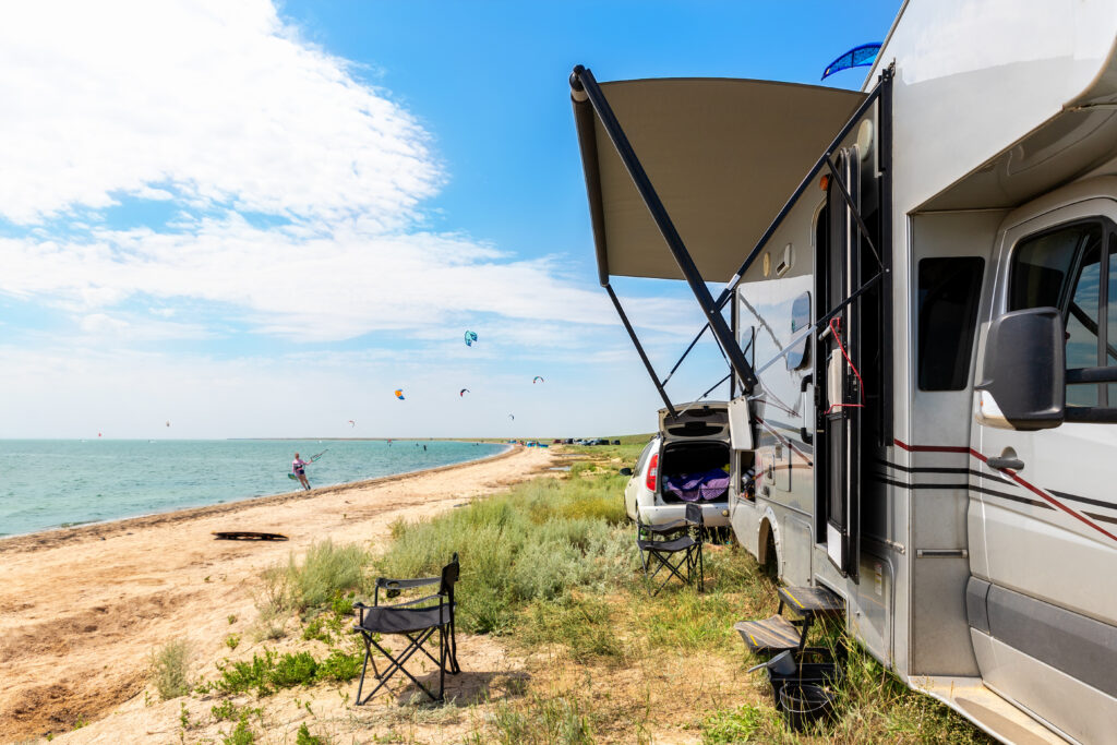 Motorhome parked beside a sandy shoreline, with camping chairs and kite surfers visible in the distance.