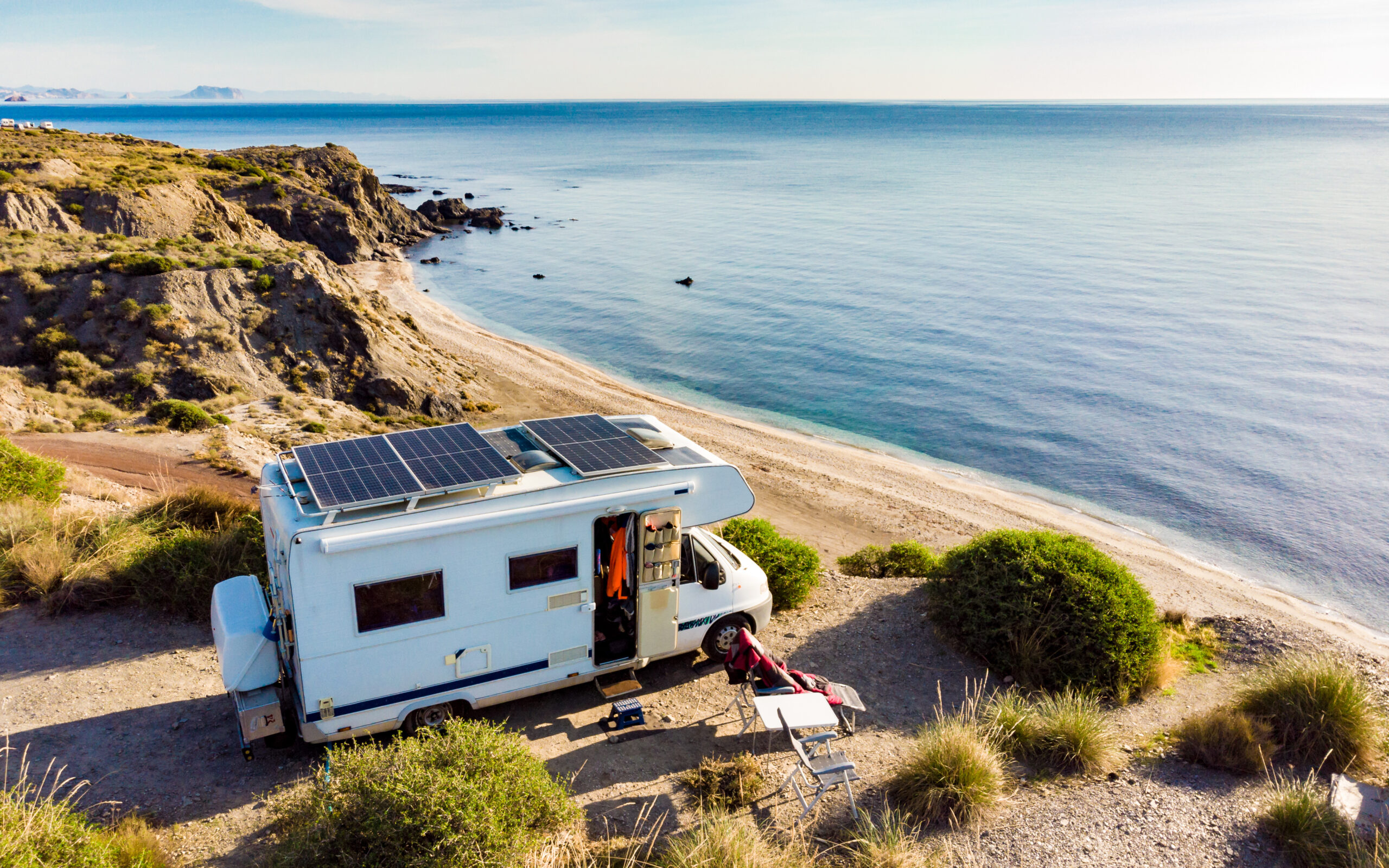 RV parked on a coastal cliff overlooking a sandy beach and calm ocean, with solar panels mounted on the roof.