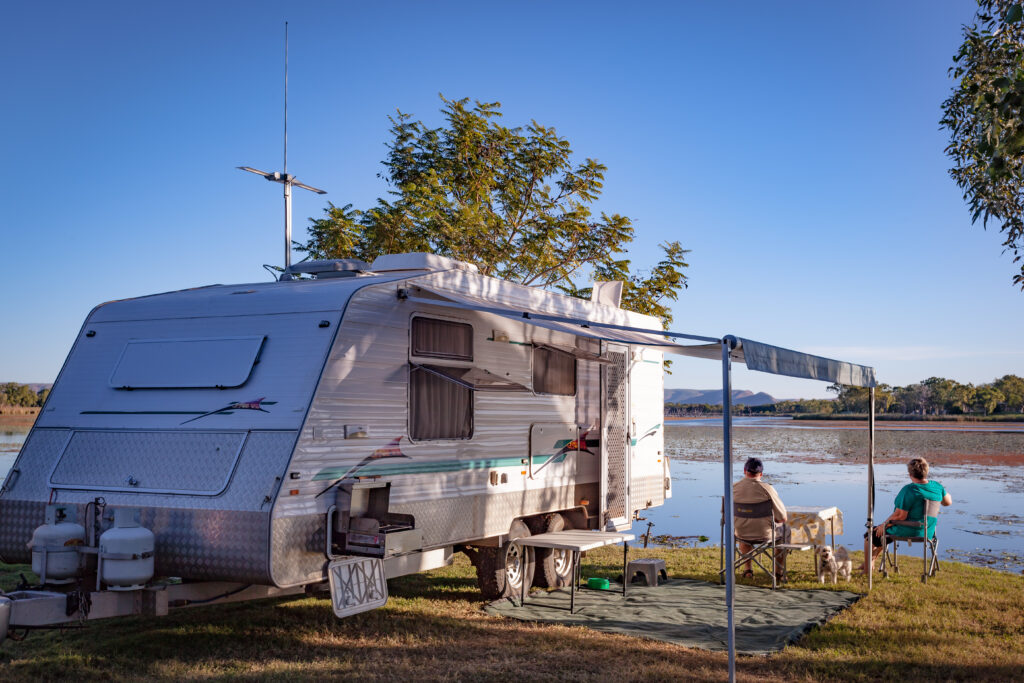 Travel trailer parked beside a lake, with two people and a dog relaxing under the awning.